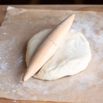 Holz-Nudelholz in einer konischen Form liegt auf einem Stück Teig auf Backpapier. Der Teig ist rund und bemehlt.